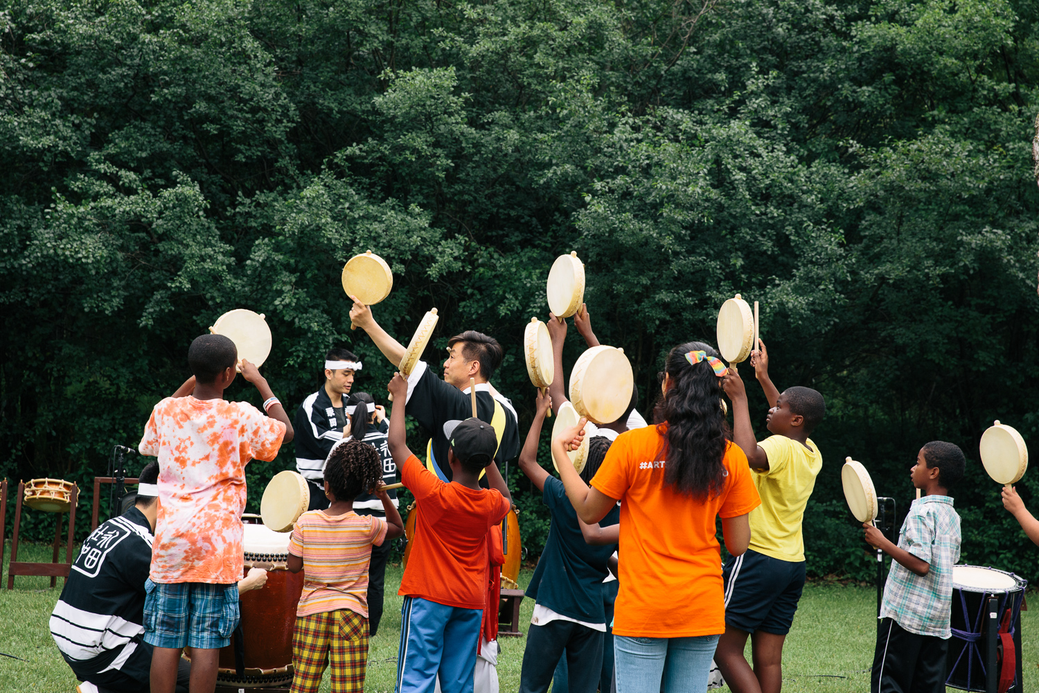 A group of children play hand drums in a field while a performer teaches them.