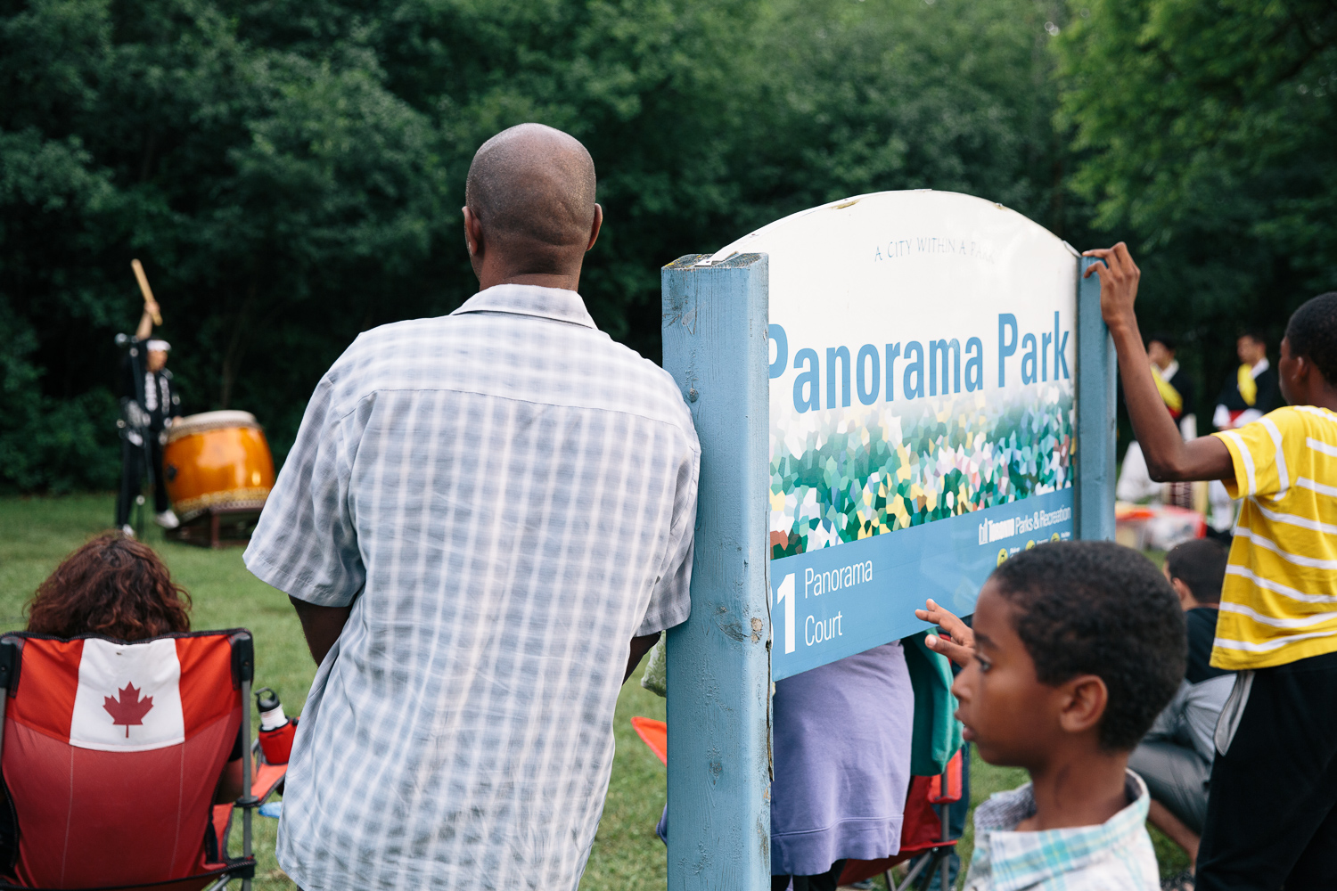 A man leans against a park sign for Panorama Park next to other audience members.