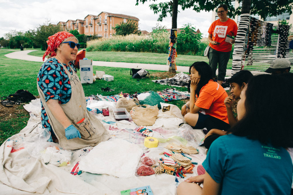 A woman in a blue and red top sits on a blanket and talks to a group of children.