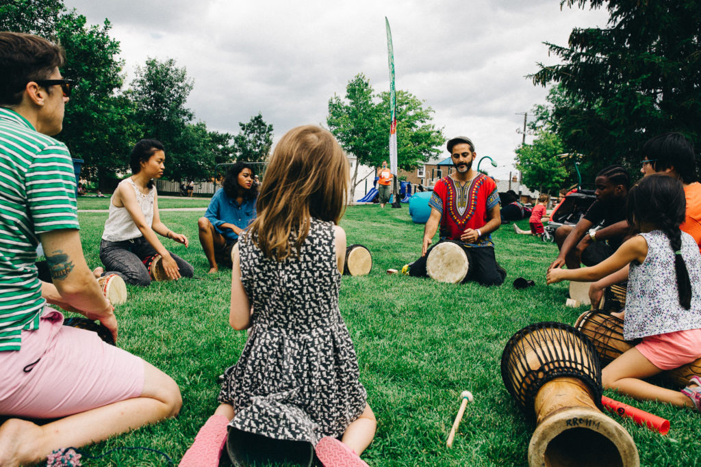 A circle of children sit on the grass, each with a drum, with an adult who is teaching them to play.