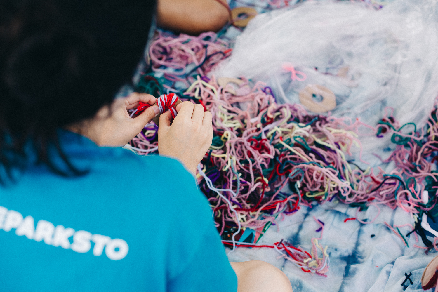 A woman in a blue shirt sits and weaves with a pile of yarn in front of her.
