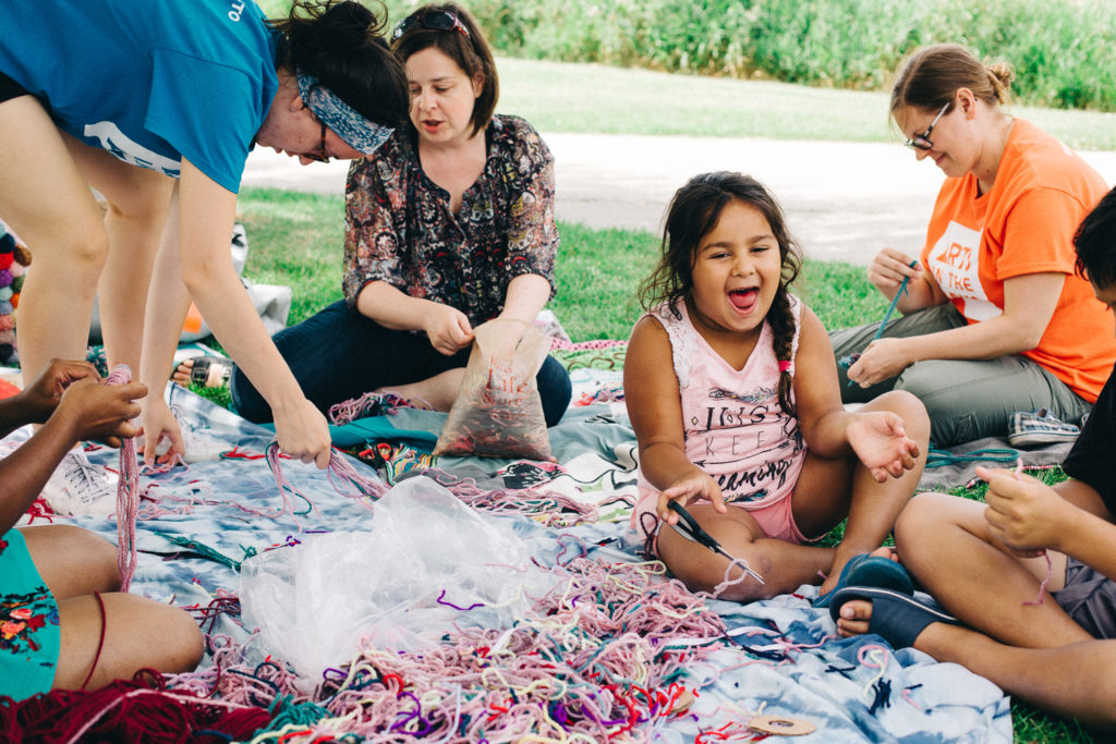 A group of people sit on a blanket and weave. A child sits with them and laughs.