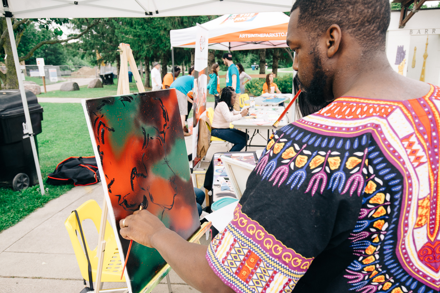 A man in a bright multicoloured shirt paints a canvas on an easel.