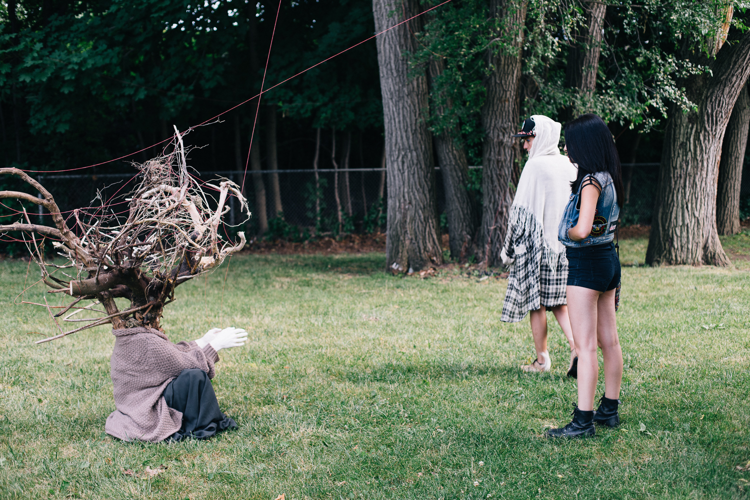 Two people stand and watch a seated person wearing a costume made of branches.