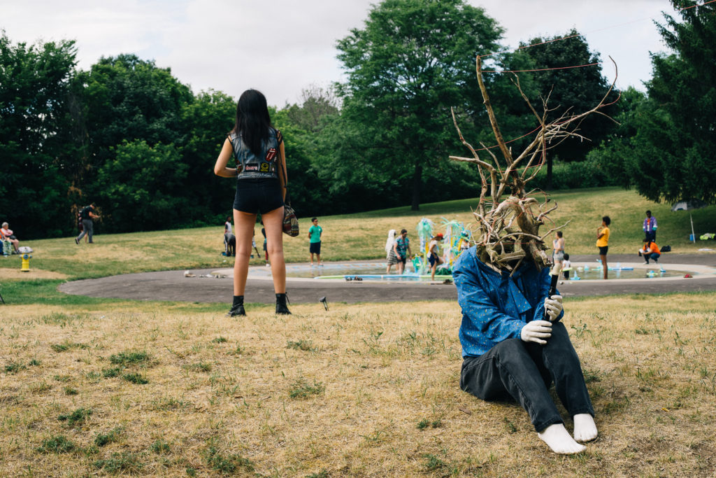 A person wearing a mask of branches sits on a grassy field near a splash pad.