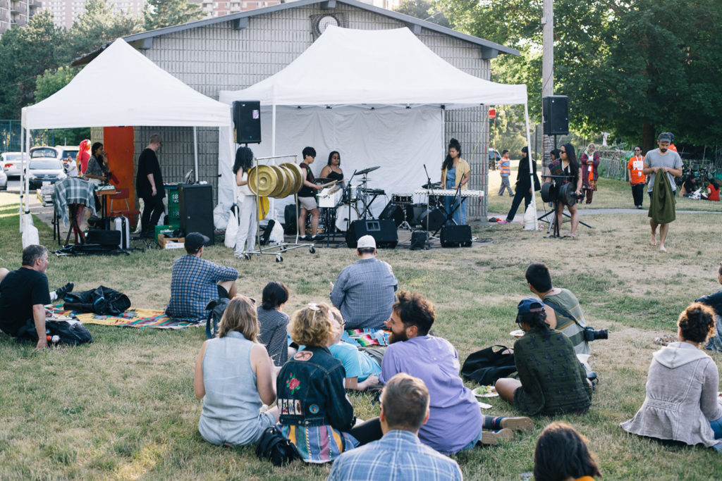 Spectators sit on a field and watch musicians perform under a tent.