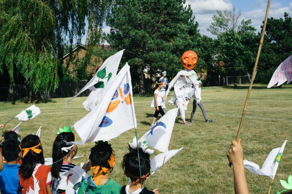 An individual wearing a large orange mask is escorted across a field as children watch.