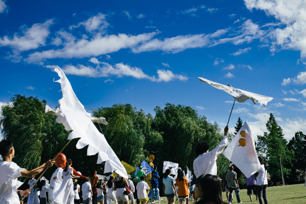 a large group of people wearing white and costumes march across a field carrying flags and large birds made of cloth.