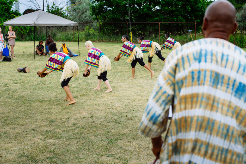 A man watches as a group of dancers in straw skirts and colourful shirts perform in a field.