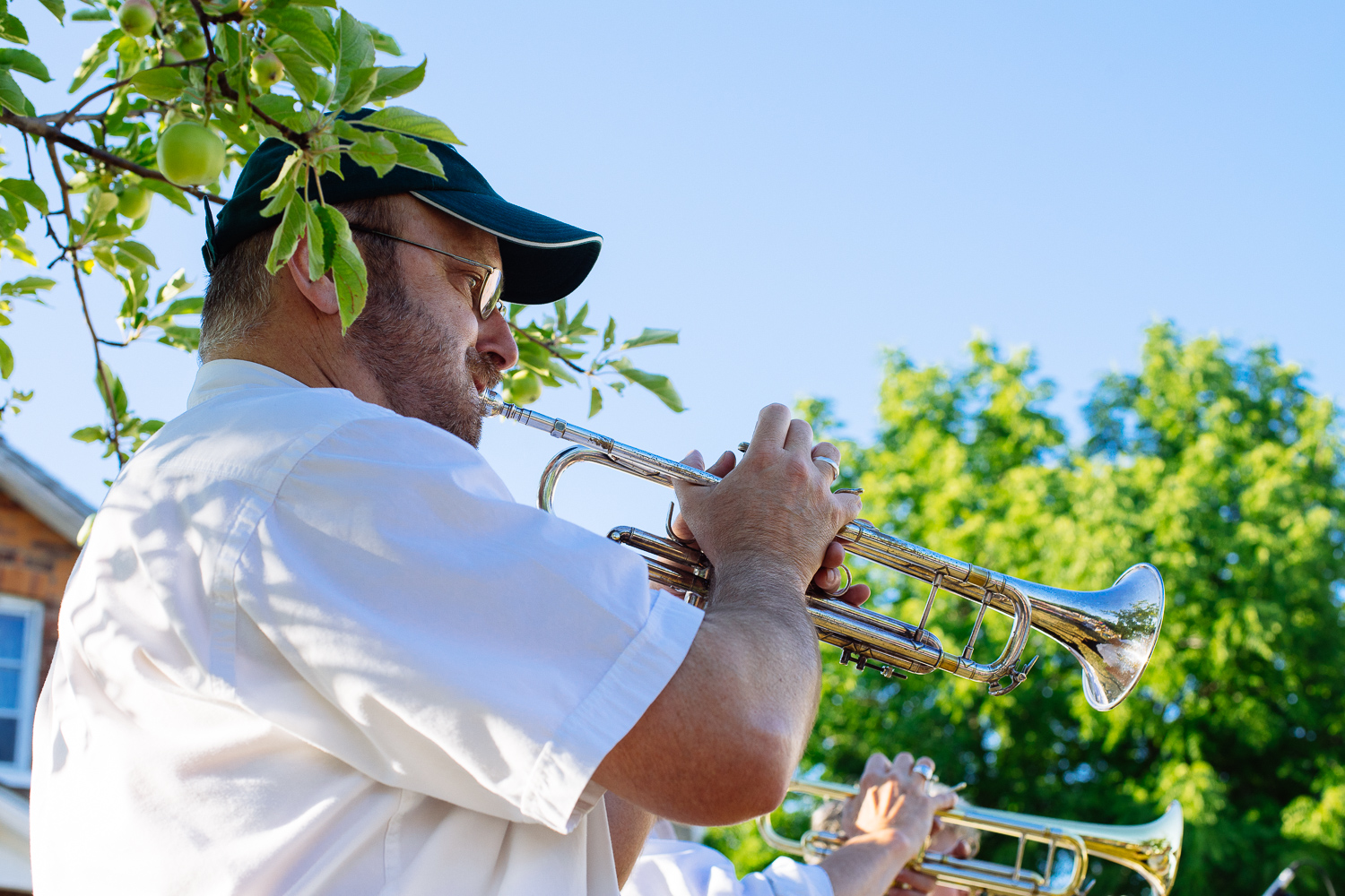 A standing man plays a trumpet under the shade of a tree.