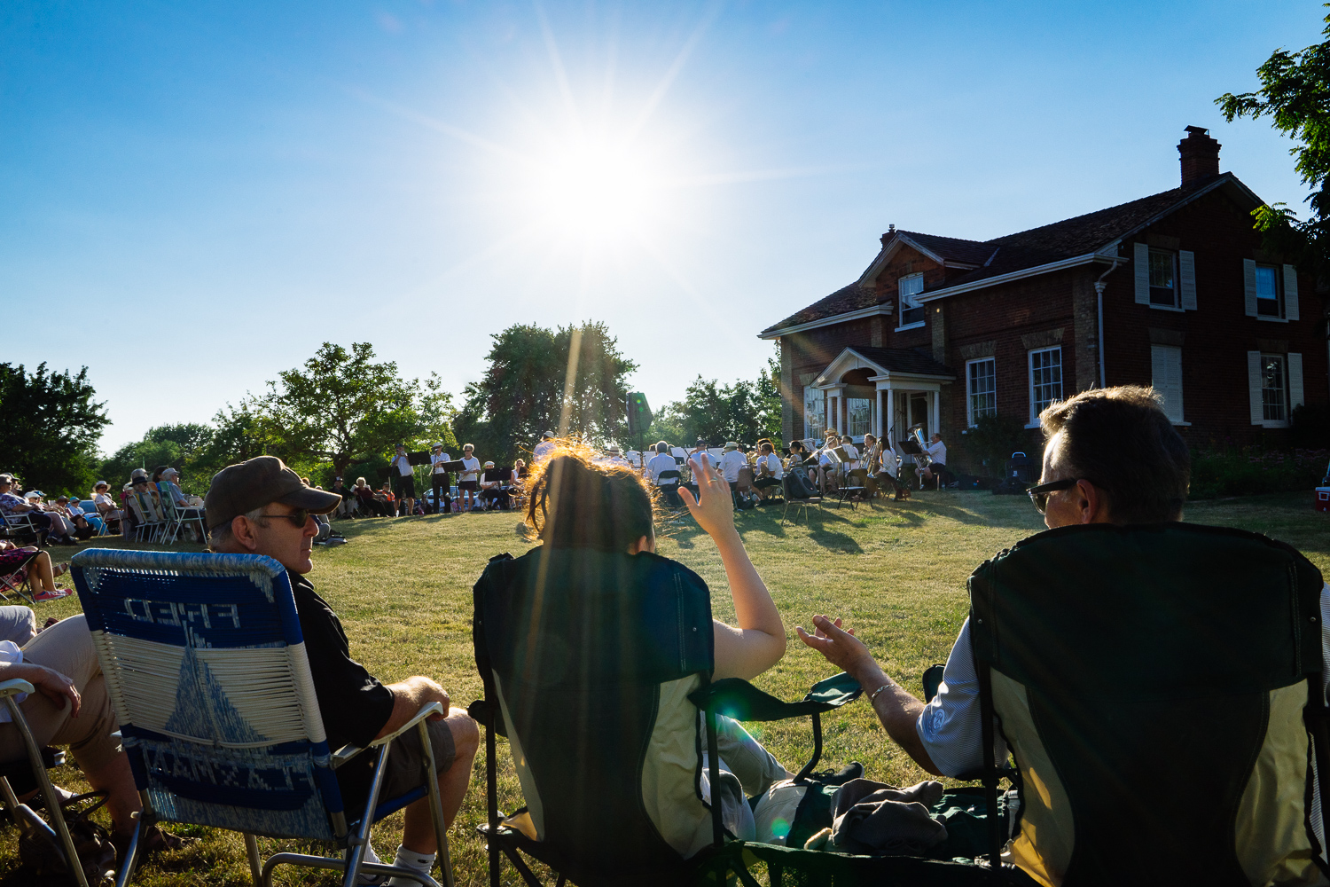 three seated audience members watch a band play in a field next to a large red brick house. The sun is shining brightly.