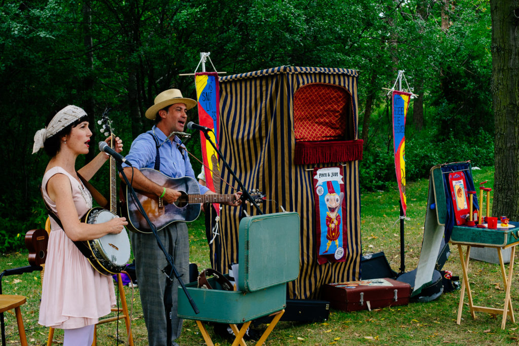 A woman and man play instruments in cosume next to a small boxed stage and two colourful flags.
