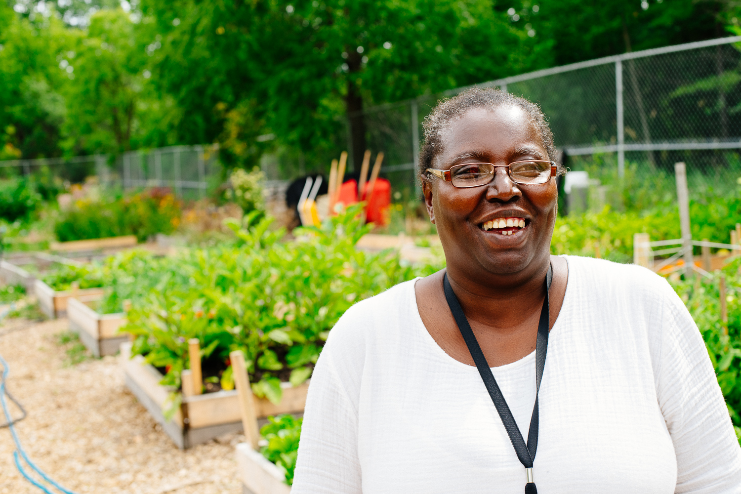 A black woman in a white shirt looks directly at the camera and smiles. Behind her is a group of vegetable gardens.