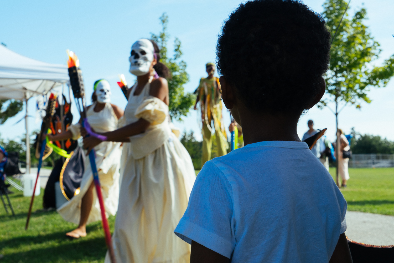 A child watches several performers in long white costumes and masks walk by.