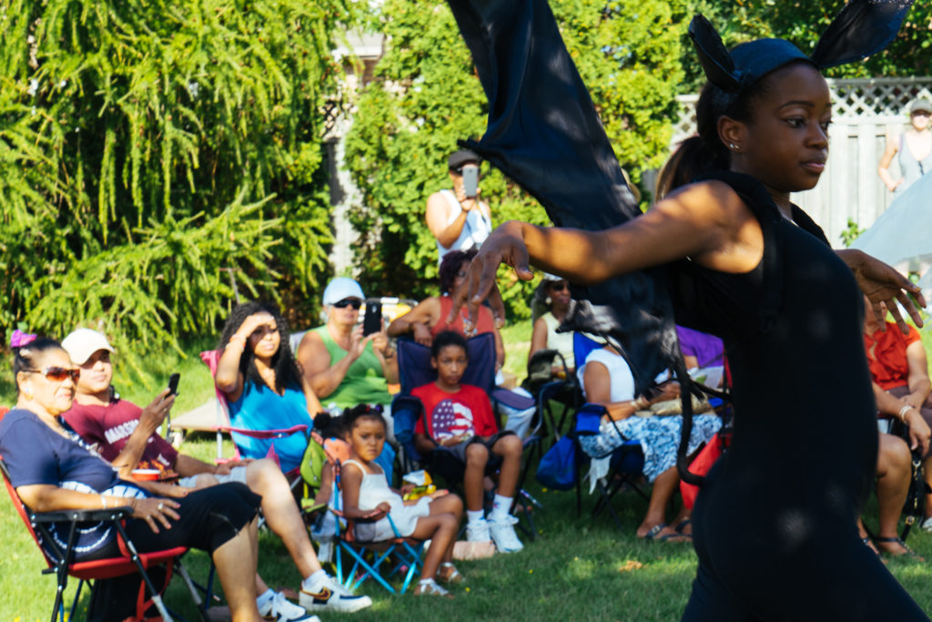 A woman in black dances as audience members are watching from the shade of a tree.