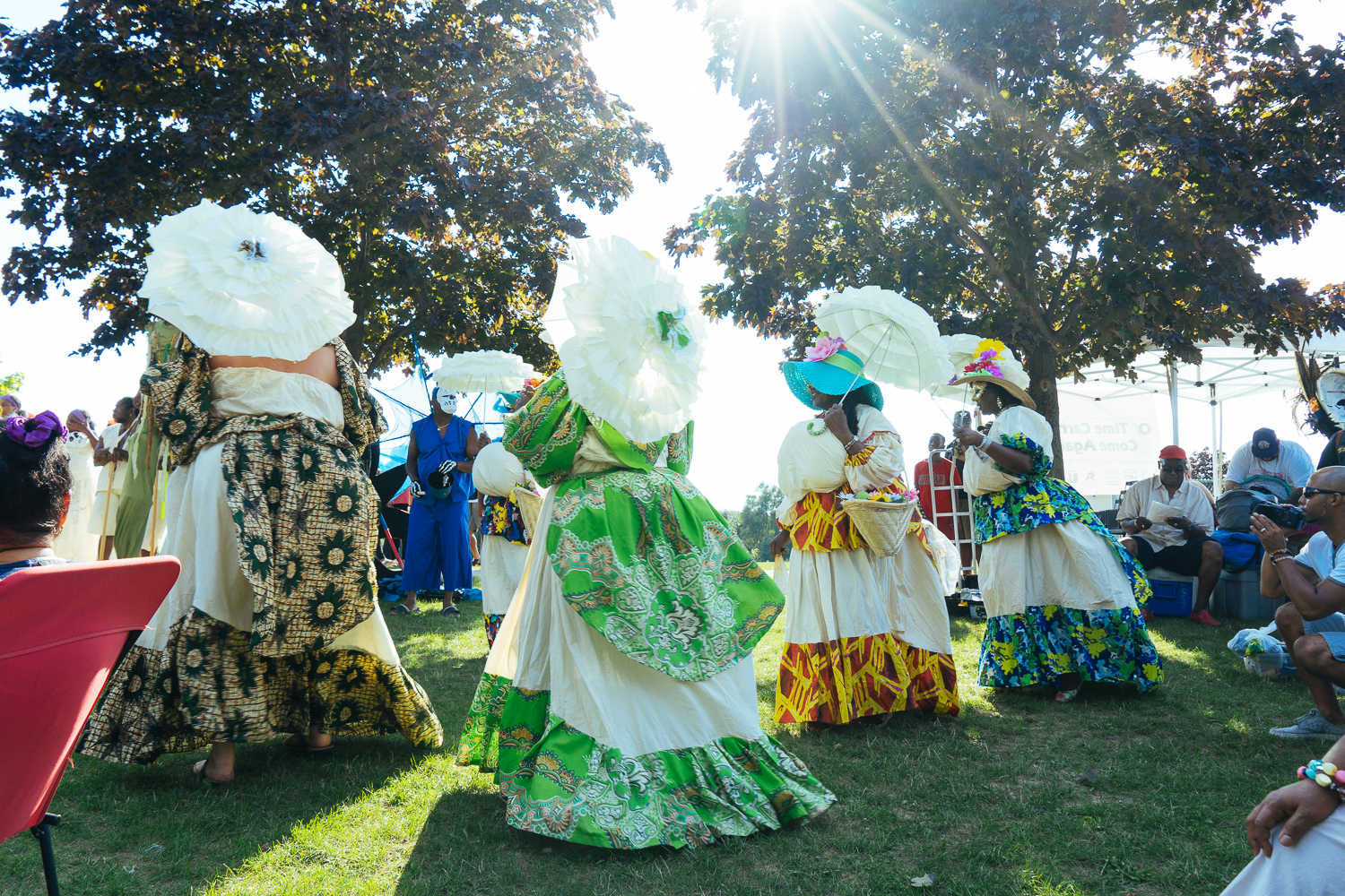 Four women in large multicoloured dresses and white parasols stand and perform in a field.