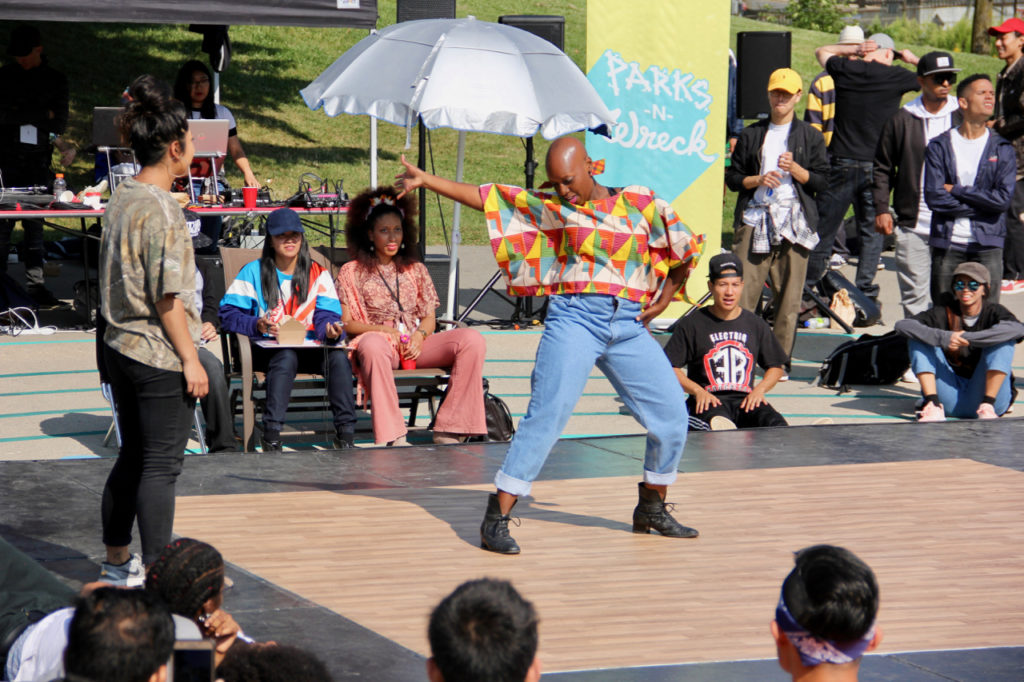 A woman in a mulicoloured shirt dances in the middle of the stage while everyone watches.