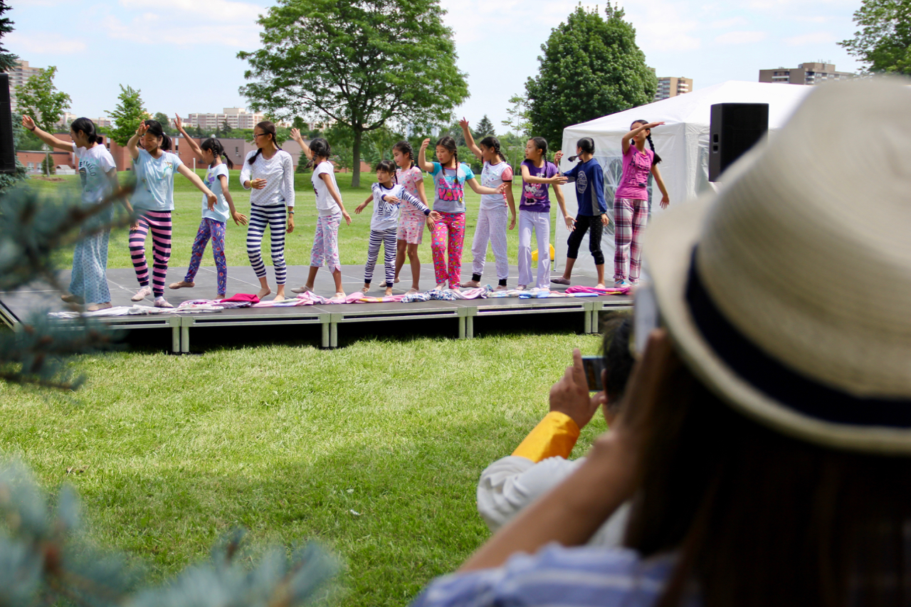 A group of children dance on stage while a woman takes a photograph.