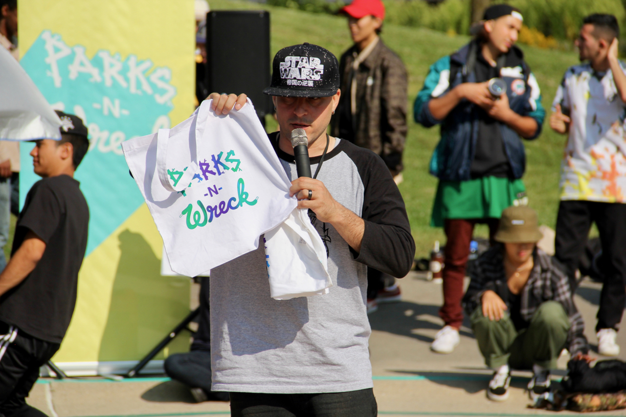 A man wth a microphone speaks to the crowd and holds a t-shirt that says "Parks N' Wreck".