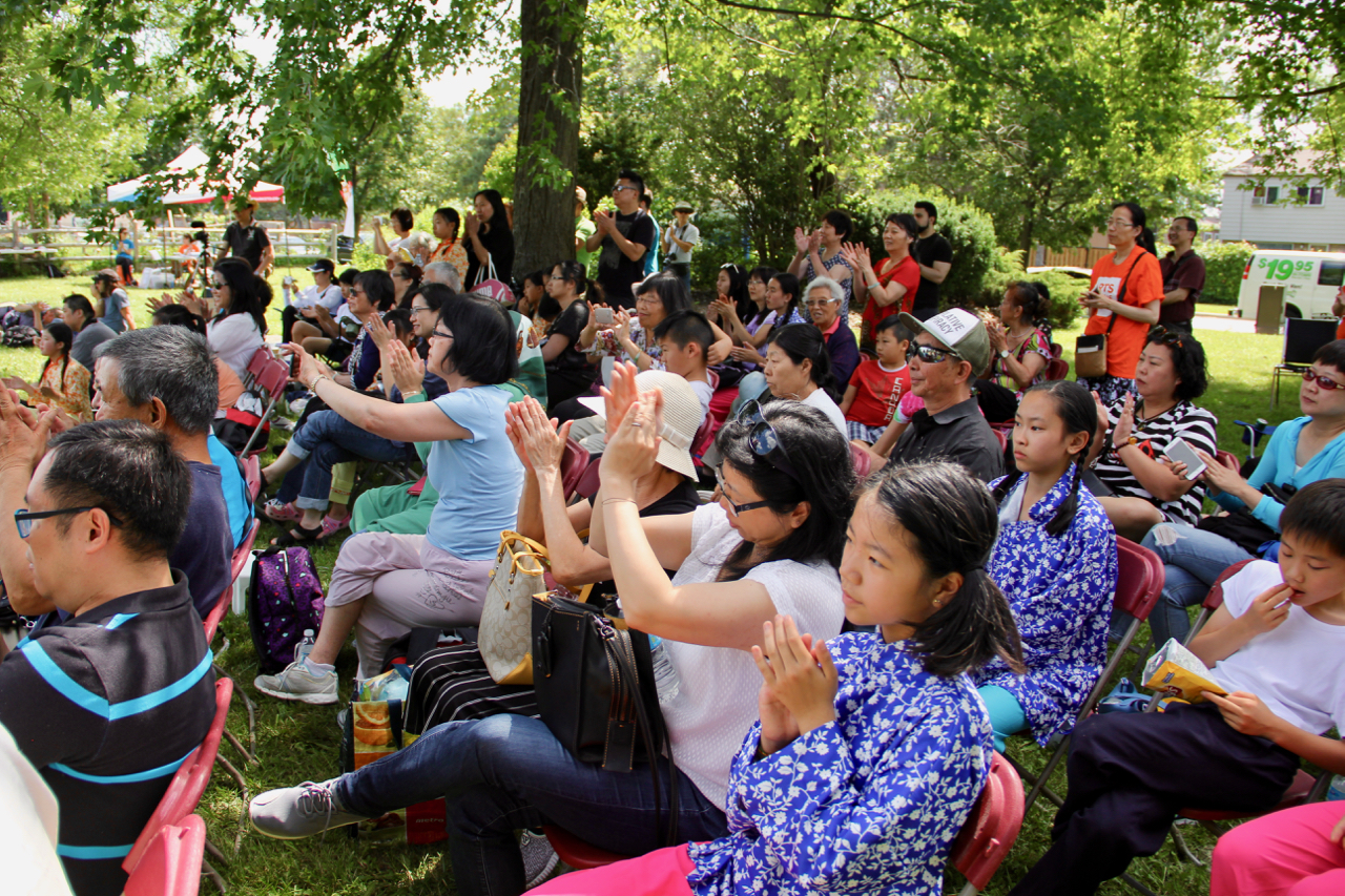 A seated crowd under the shade of the tree claps for a performance.
