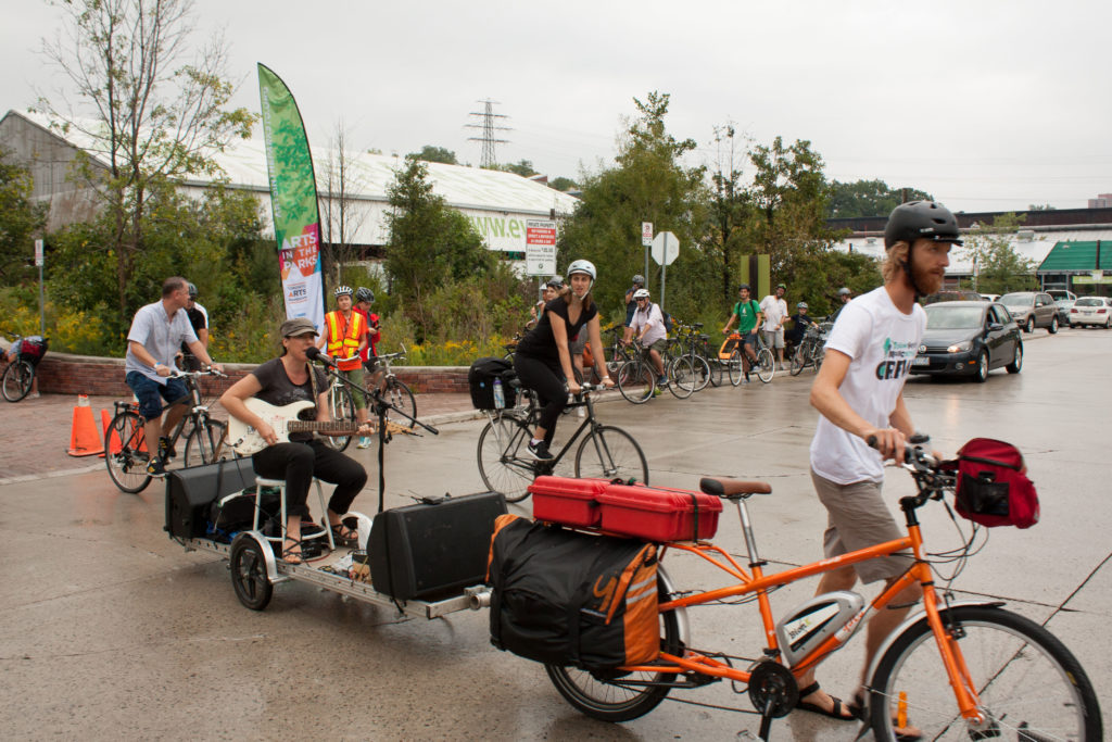 Image description: a musician and equipment are pulled by bikes across a road.