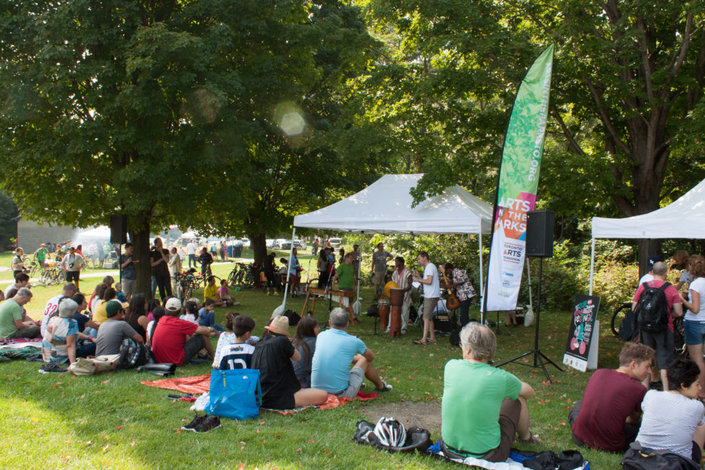 Image description: a group of people on blankets watch a band perform under a tent near trees.