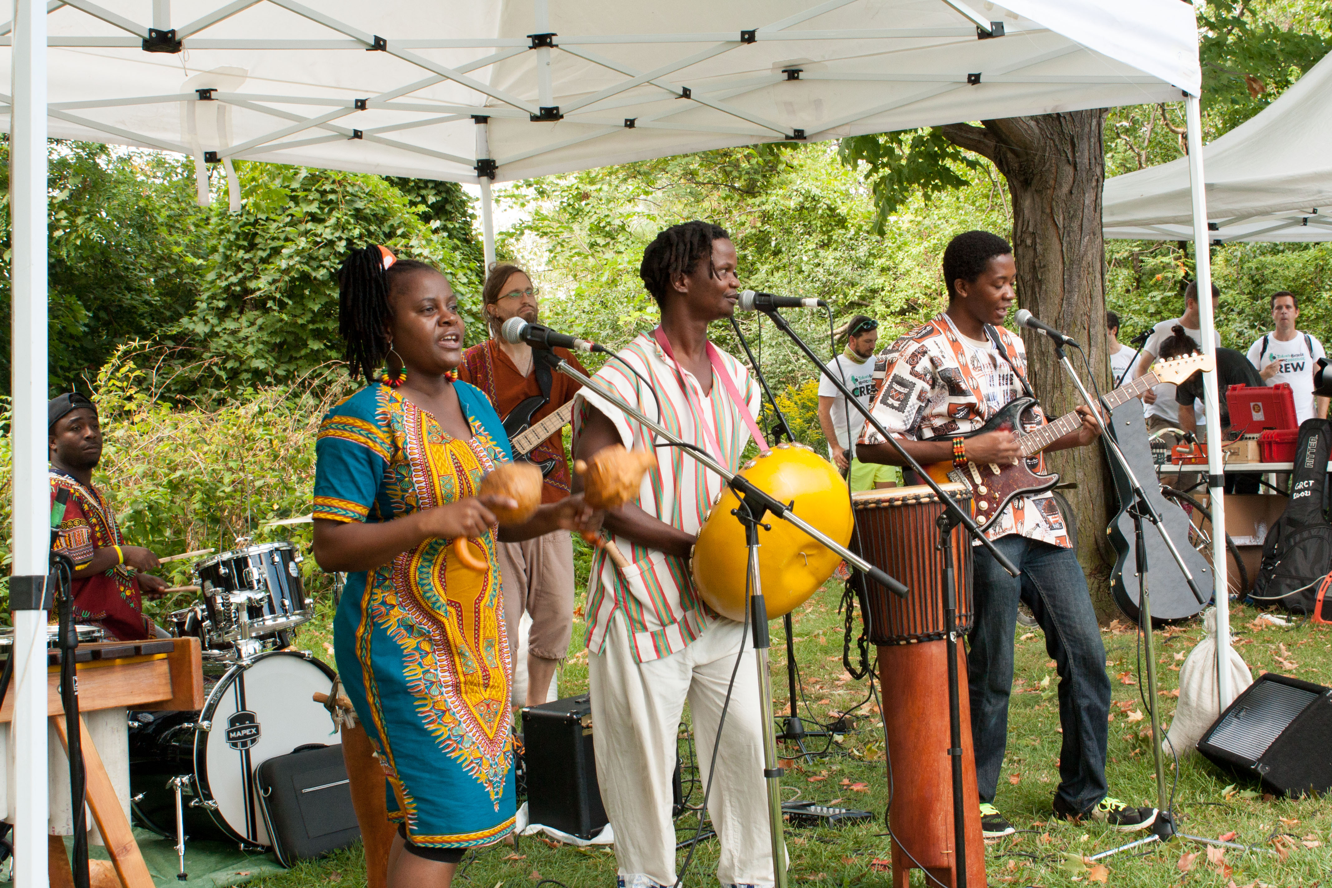 Image description: three musicians play percussion instruments and sing under a tent.