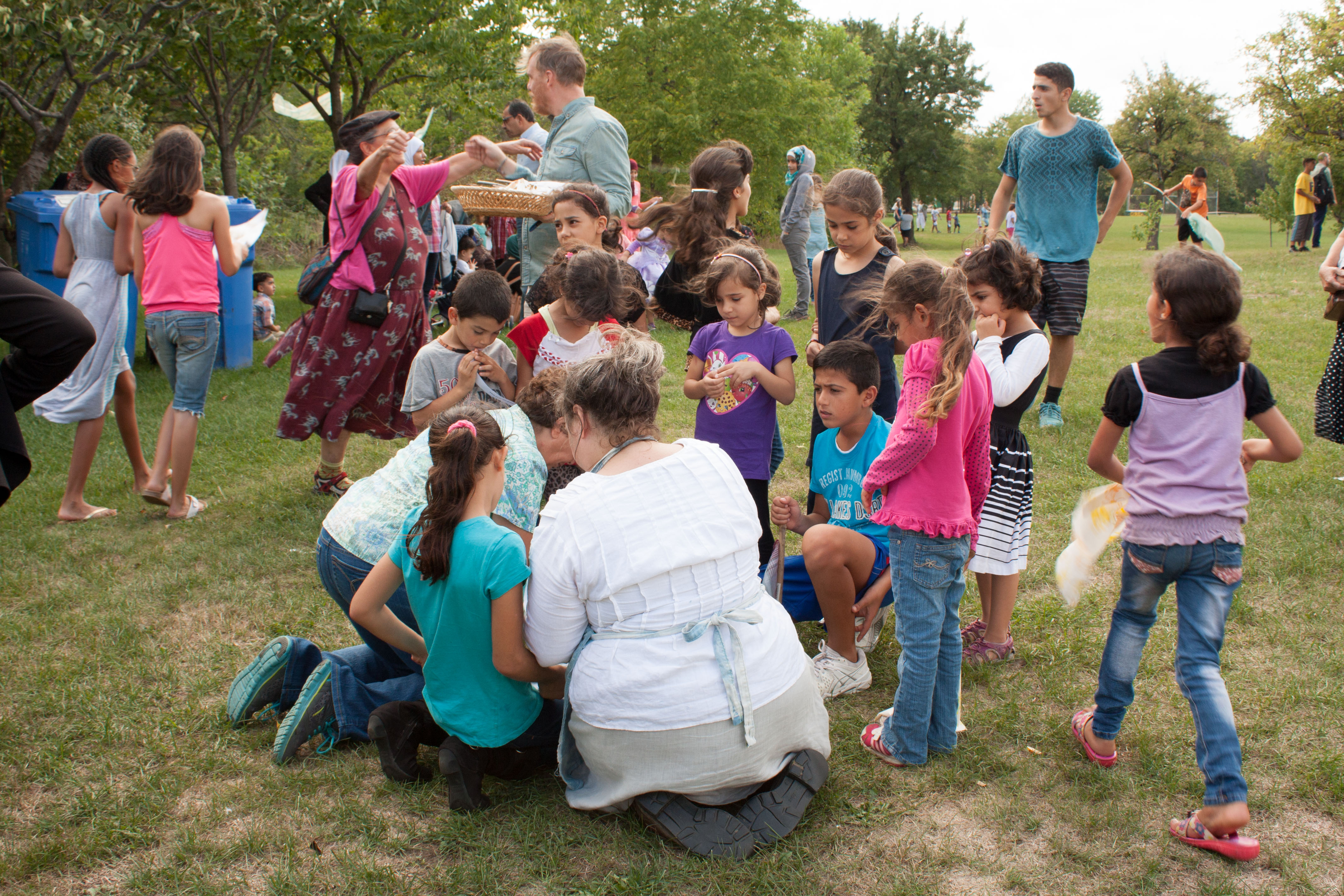 An adult sits on the grass and is surrounded by a group of children looking on.