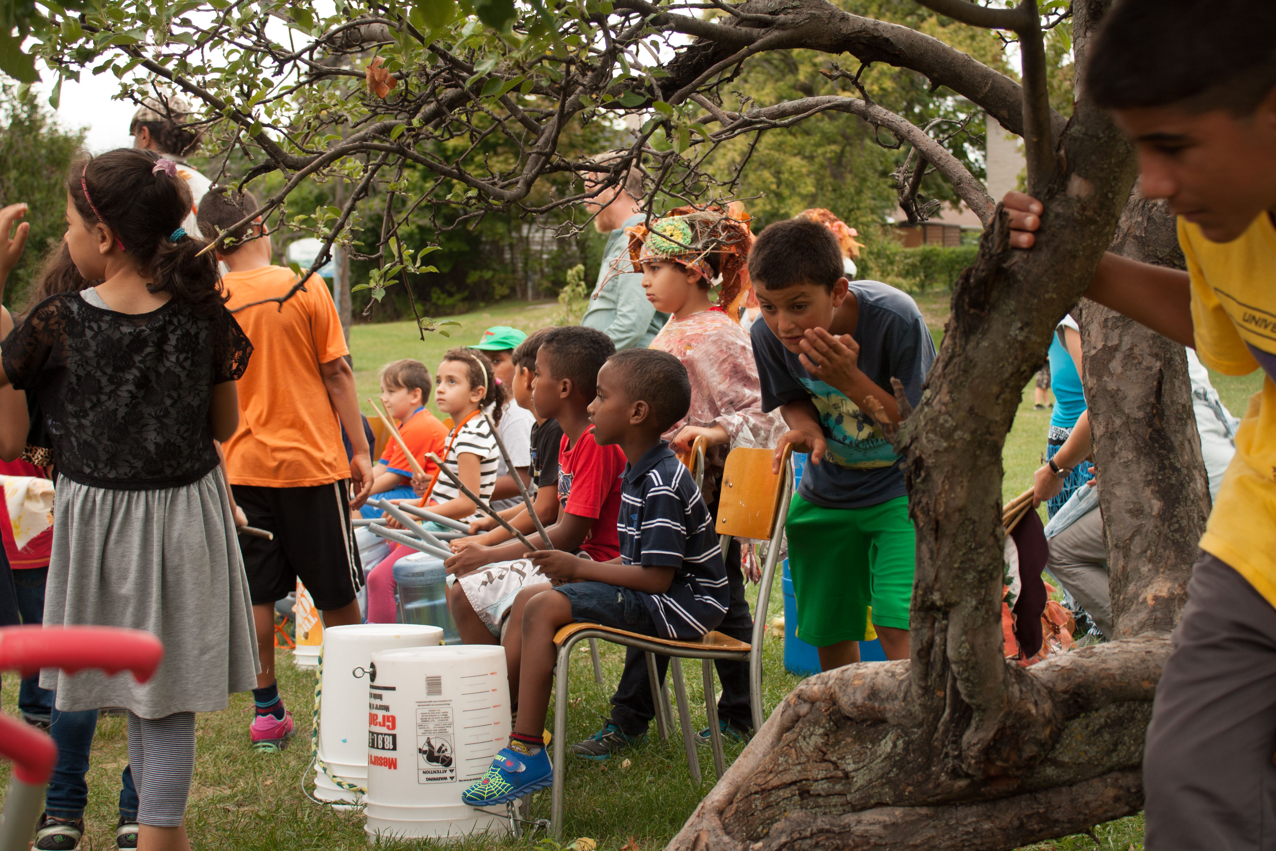 A group of children play buckets as drums under a tree.