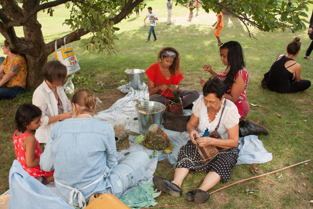 Adults and children sit on blankets under a tree working on crafts together.