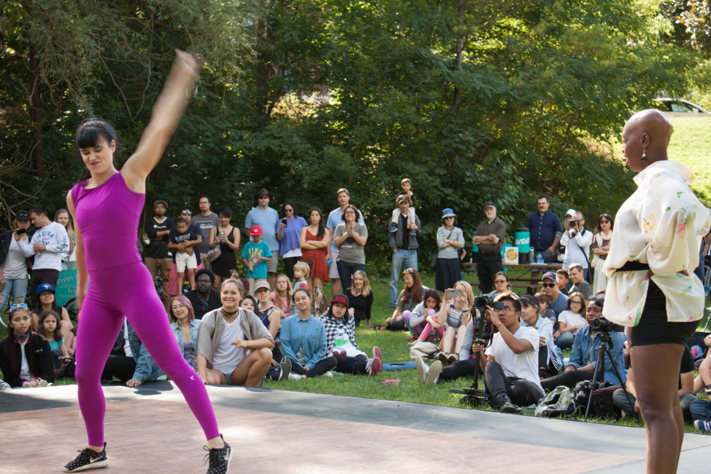 Two performers dance on a raised stage while a large group of people sit and watch.