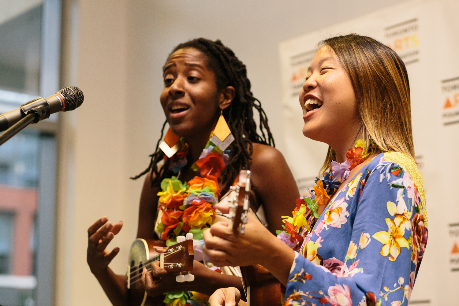 Two women stand in front of a microphone. They are singing and playing ukeleles.