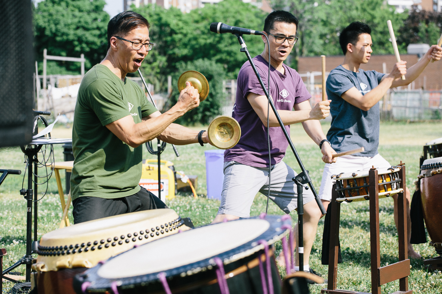Three men play drums and cymbals as they perform on the grass.