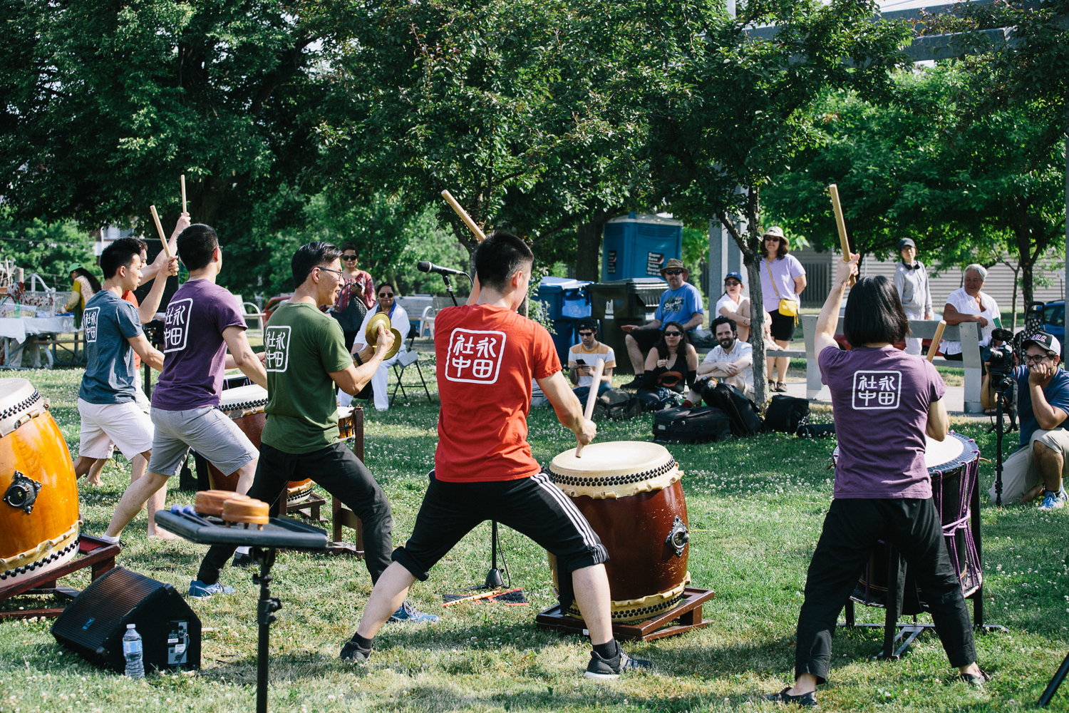 A group of performers in colourful shirts play the drums in a field as audience members watch from the shade of the trees.