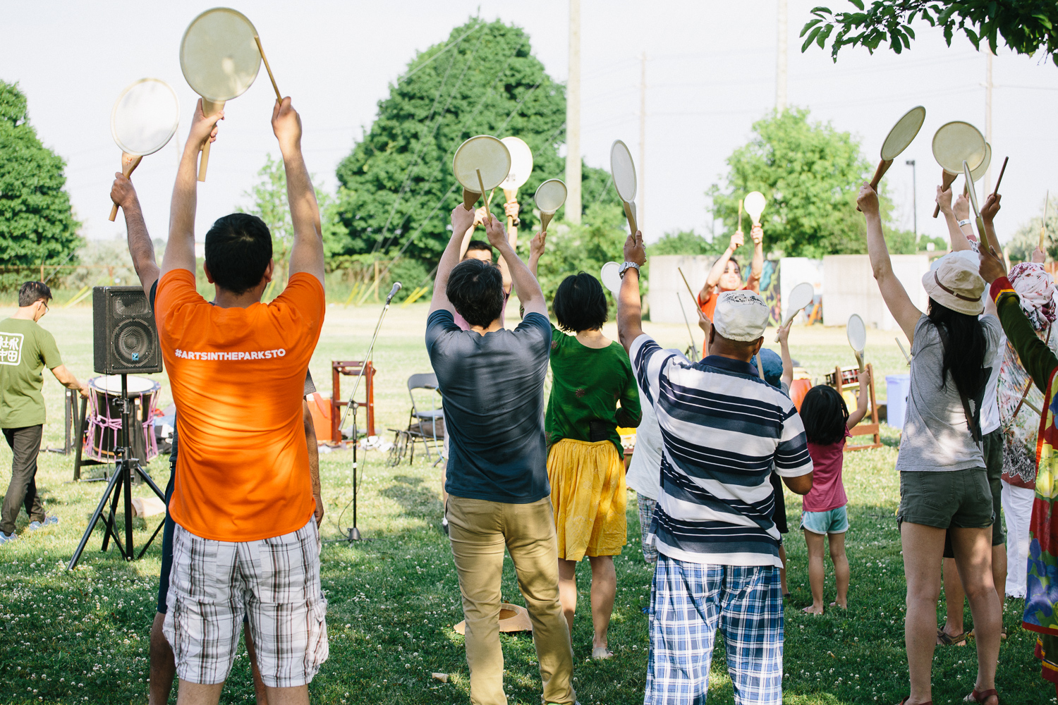 A group of audience members stand and raise hand drums above their head as they participate in a workshop on the grass.