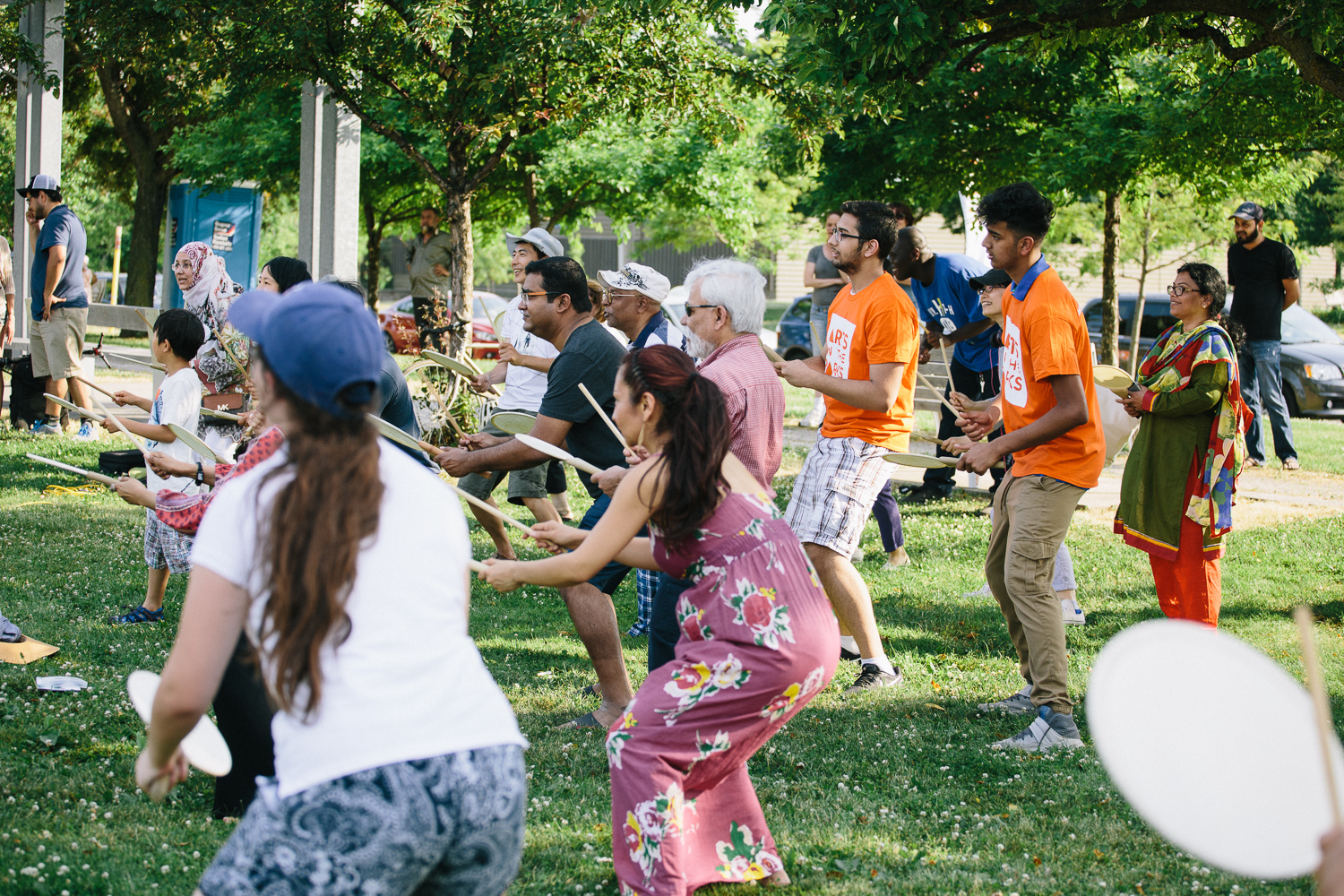 A group of audience members stand and play hand drums as they participate in a workshop on the grass.