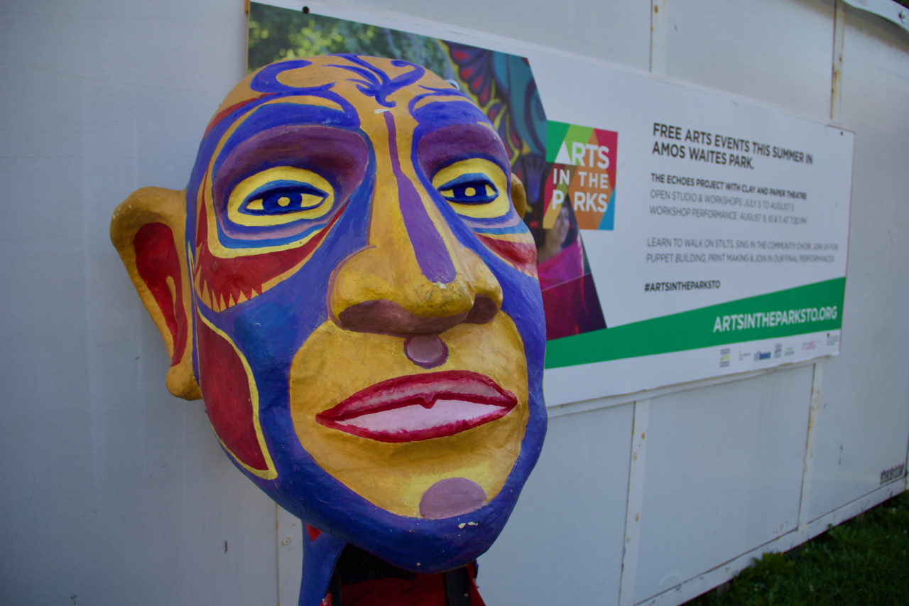 A large, multicoloured paper mache mask sits next two a large metal storage container with a banner on it.