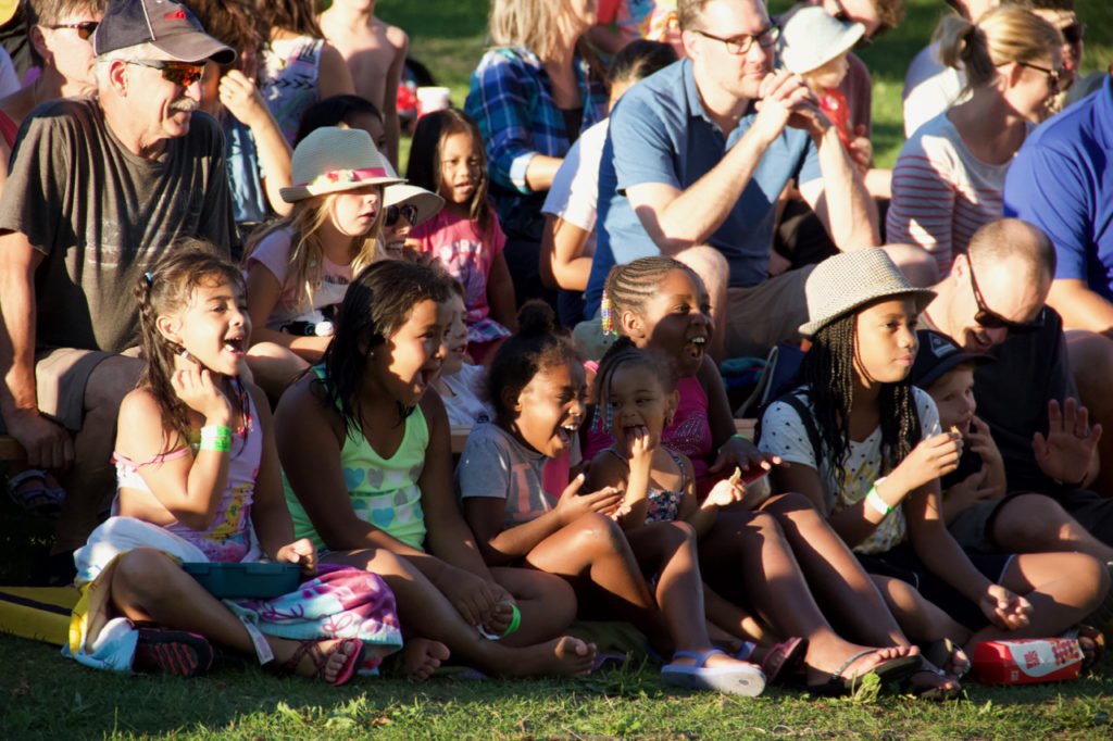 A large crowd of children are seated on the grass and clapping as they watch a performance.
