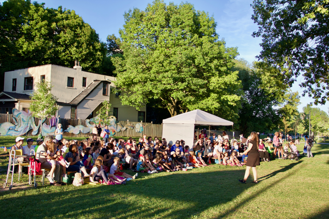 A large crowd of children and adults are seated and watching a performer in the middle of a field.