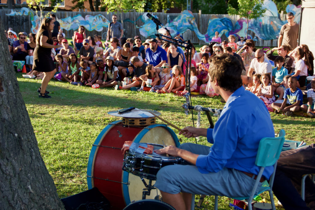 A man sits and plays the drums and watches a crowed seated to his right.