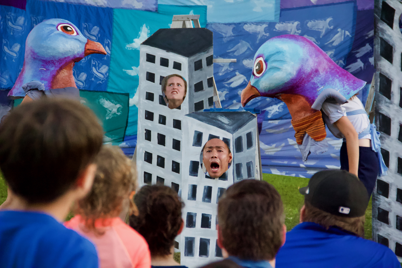Two performers with large paper mache pigeon heads stand on either side of a painted building with two performers sticking their heads through face holes.