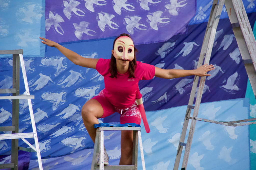 A woman wearing a white mask perches on top of a ladder in front of a multicoloured backdrop