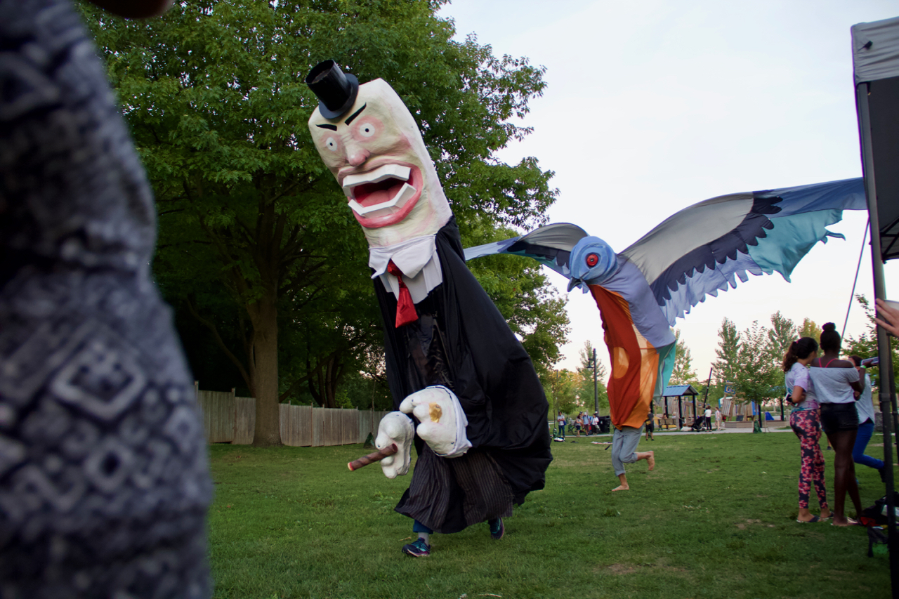 A giant puppet in a black suit runs through a field and is followed by a giant bird puppet handled by three performers.