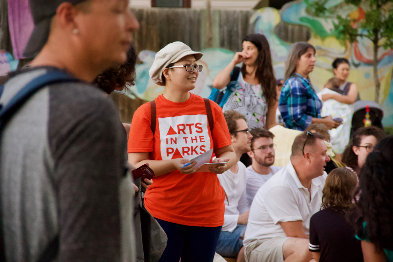 A volunteer walks through the crowd. She is wearing an orange shift that says Arts in the Parks.