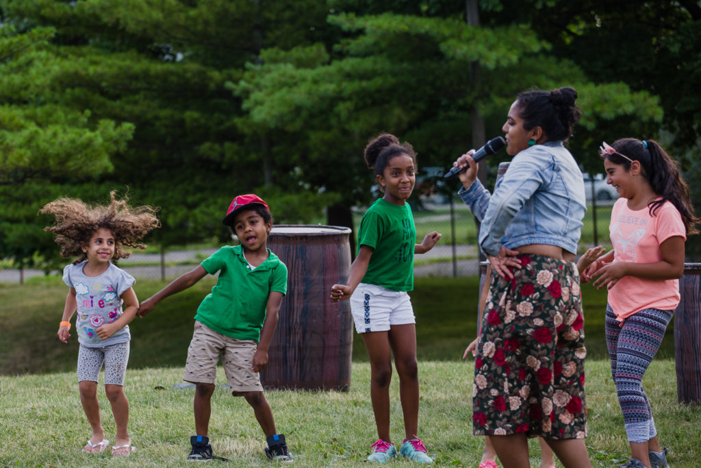 A woman in a jean jacket and flowered skirt sings as a group of children dance around her on the grass.