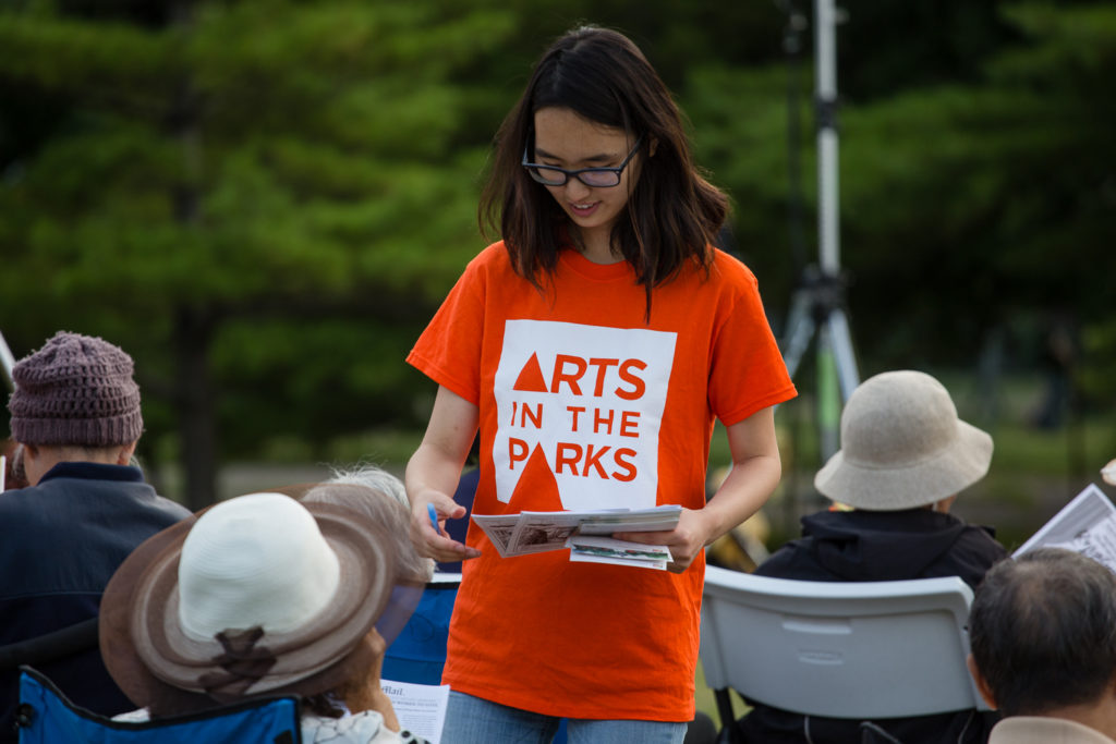 A volunteer passes out pieces of paper. She is wearing an orange shirt that says "Arts in the Parks".