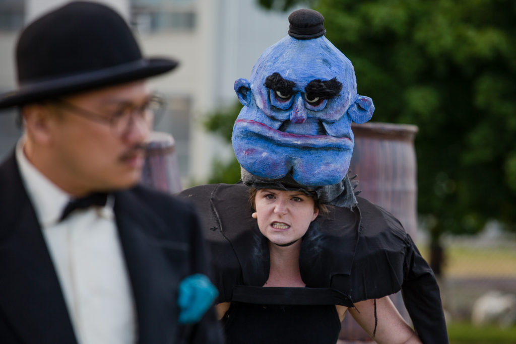 A woman wears a large blue mask on the top of her head. She stands behind a man in a black suit and black hat.
