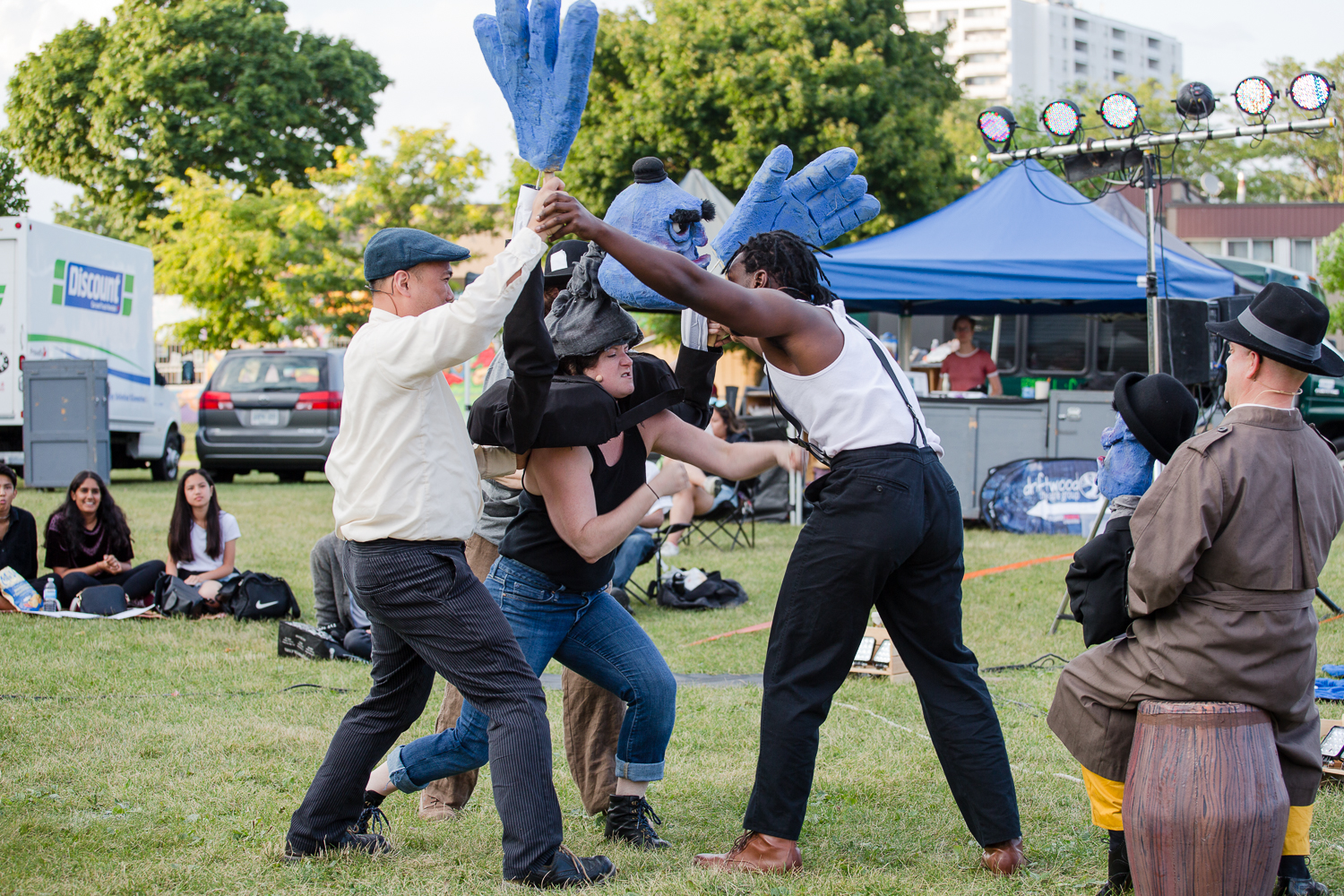 Three performers stage a fight. Two men hold the woman performer wearing a blue mask and giant blue foam hands.