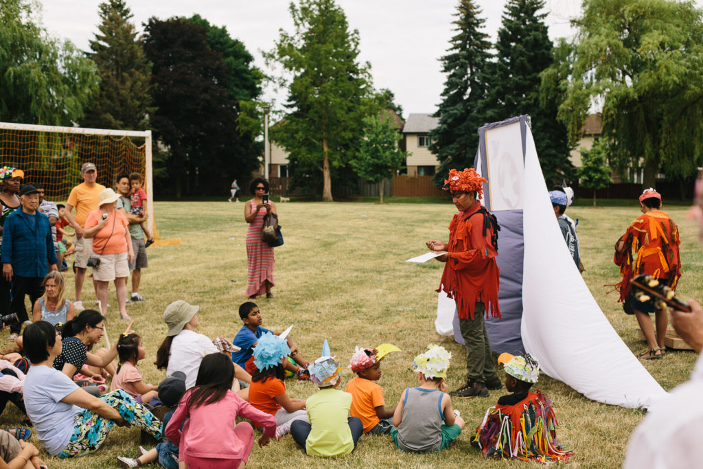 Two performers in costumes stand in front of a white backdrop and speak to a crowd of children.