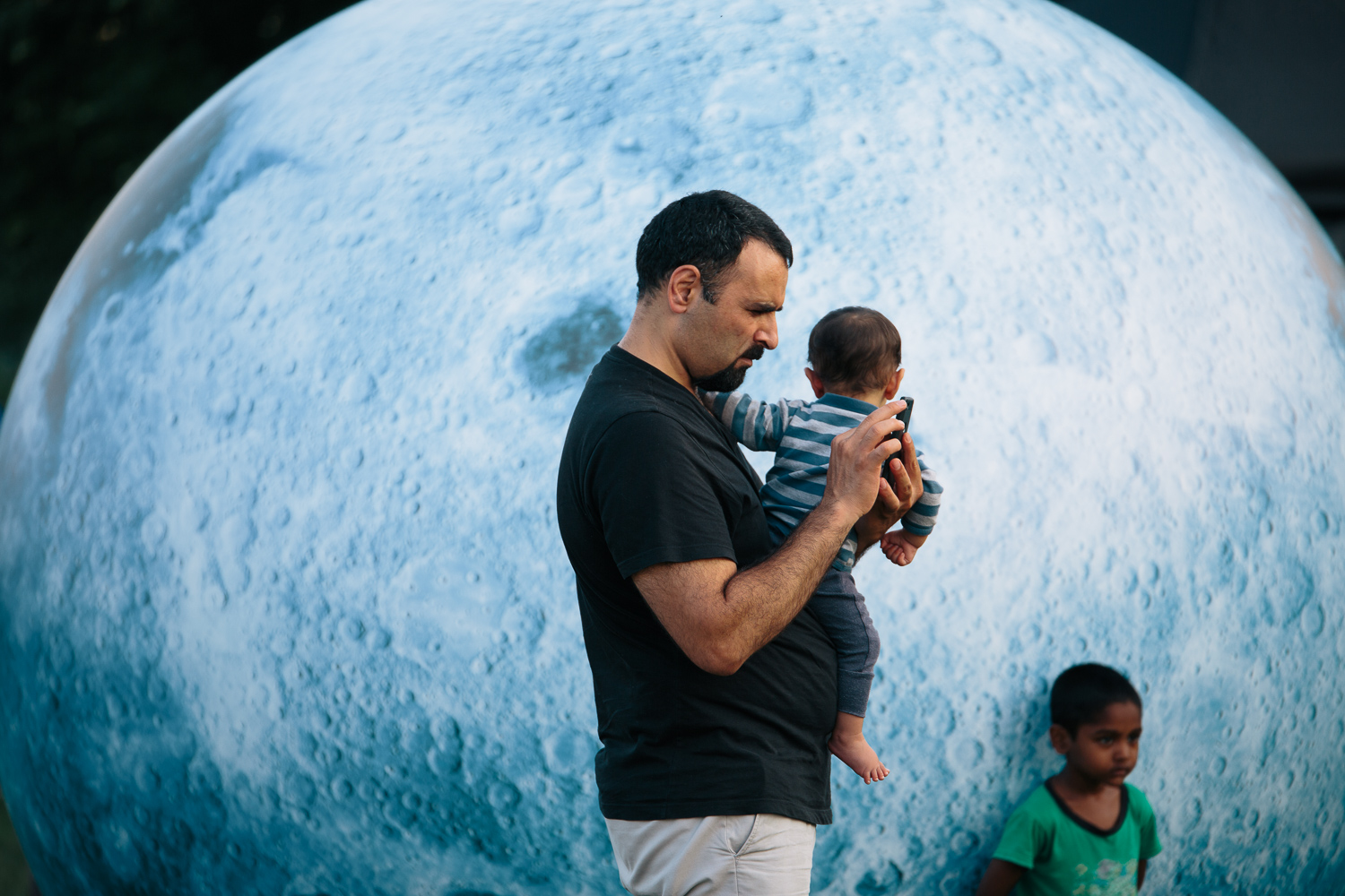 A man holds a child and stand in front of a large blue sphere.